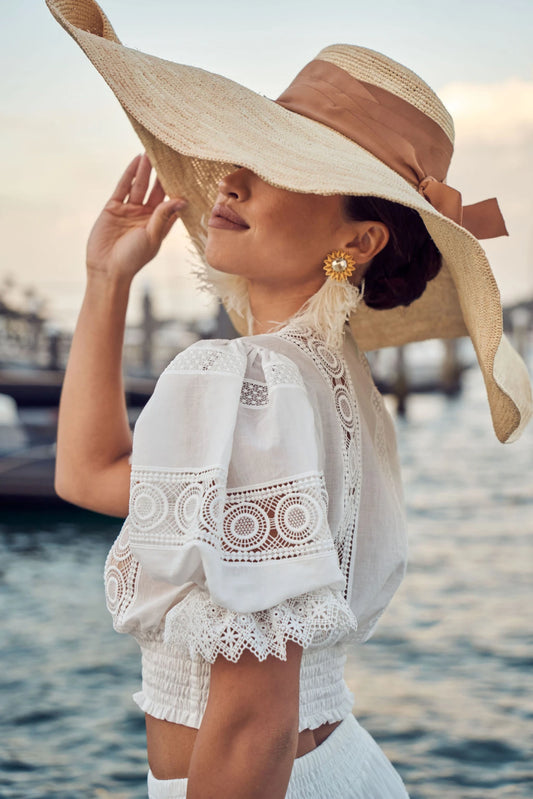 Model in white embroidered blouse wearing the hat pulled low over her face; styled for seaside sophistication.