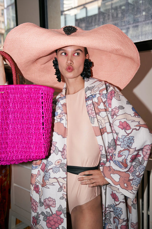 Model wearing the Lady Ibiza Hat in a playful editorial setting, paired with a floral kimono, pink bodysuit, and oversized woven fuchsia tote—exuding bold, eclectic summer glamour.