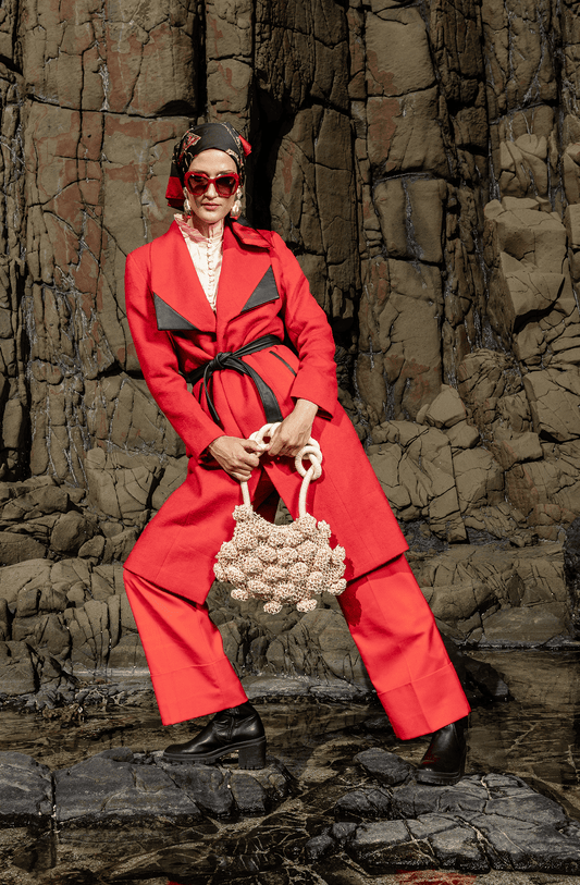 Model in red outfit seated against a Peruvian stone backdrop holding the cream Ameba Mesh Bag, highlighting its sculptural form and artisanal texture.