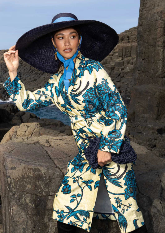 Model dressed in a printed co-ord set and wide-brim hat holding the Sapphire Moon Clutch against a rocky coastal backdrop.