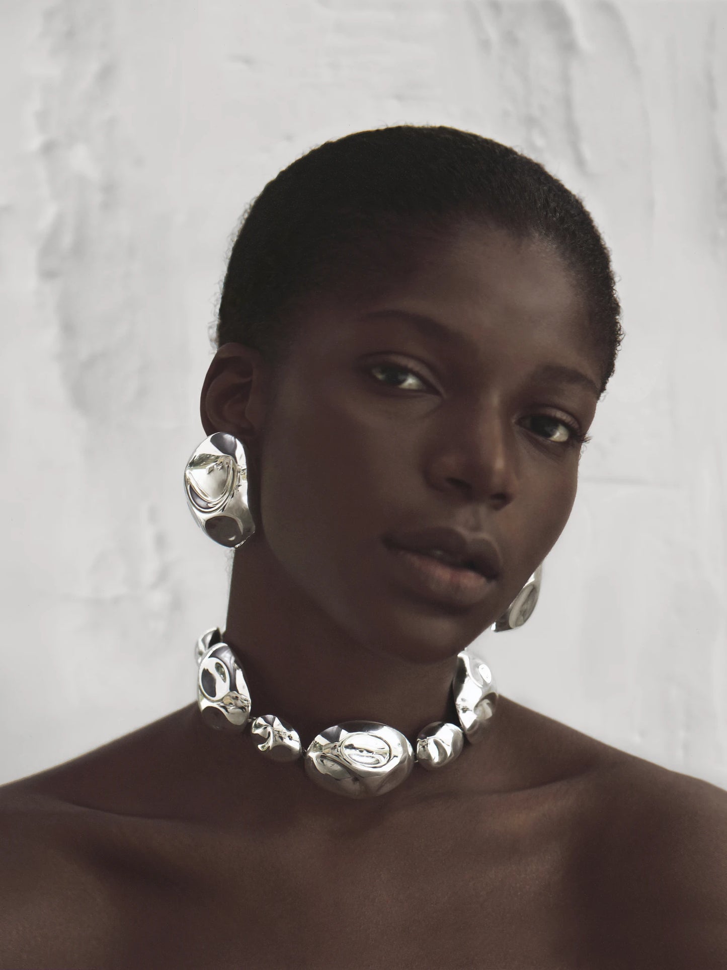 Model with cropped hairstyle showcases silver statement earrings paired with a coordinating silver sculptural necklace, exuding bold elegance.