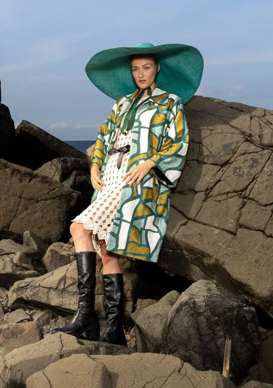 Front of the Stained Glass Coat with structured silhouette and vibrant green-gold print, photographed in Miami, Florida – heritage fashion reimagined by My Paloma. Wearing a big green hat standing on a rocky shore line
