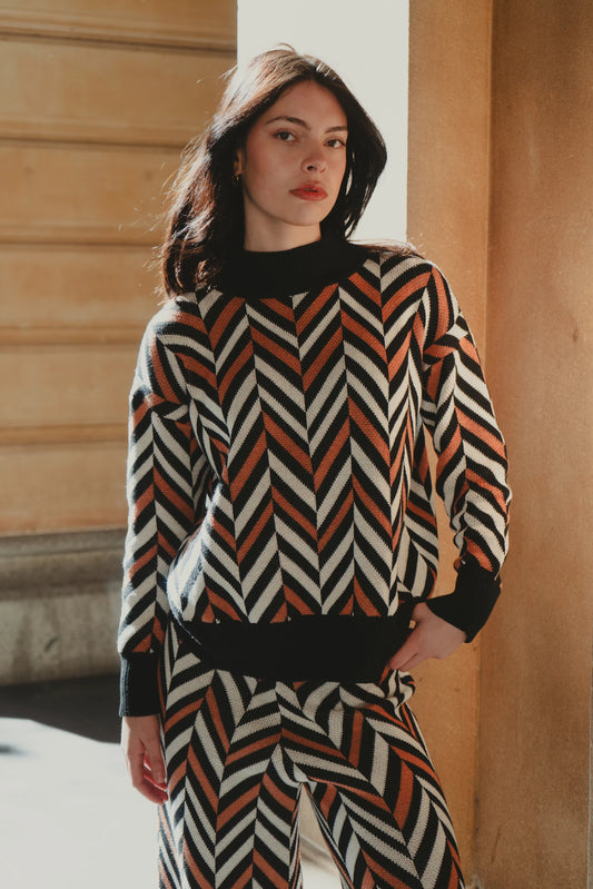 Model leaning against an arched stone pillar, wearing the My Paloma Zig Zag co-ord set with wide-leg pants and chevron print, styled with ankle boots for an elevated streetwear aesthetic in a luxury urban setting.