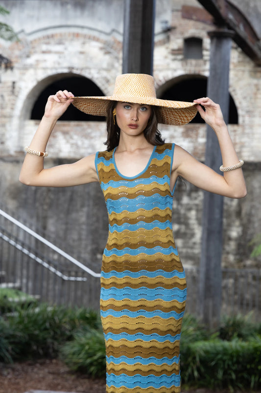 Model wearing the Chalan Sunhat paired with a wave-knit dress, holding the brim confidently while standing in front of rustic stone architecture—blending artisanal fashion with European-inspired elegance.