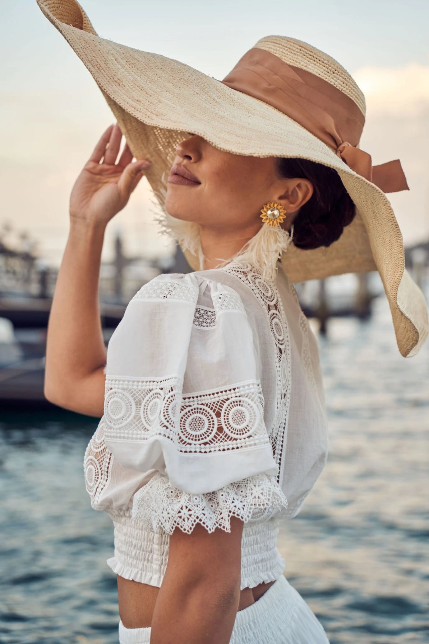 Model in white embroidered blouse wearing the hat pulled low over her face; styled for seaside sophistication.