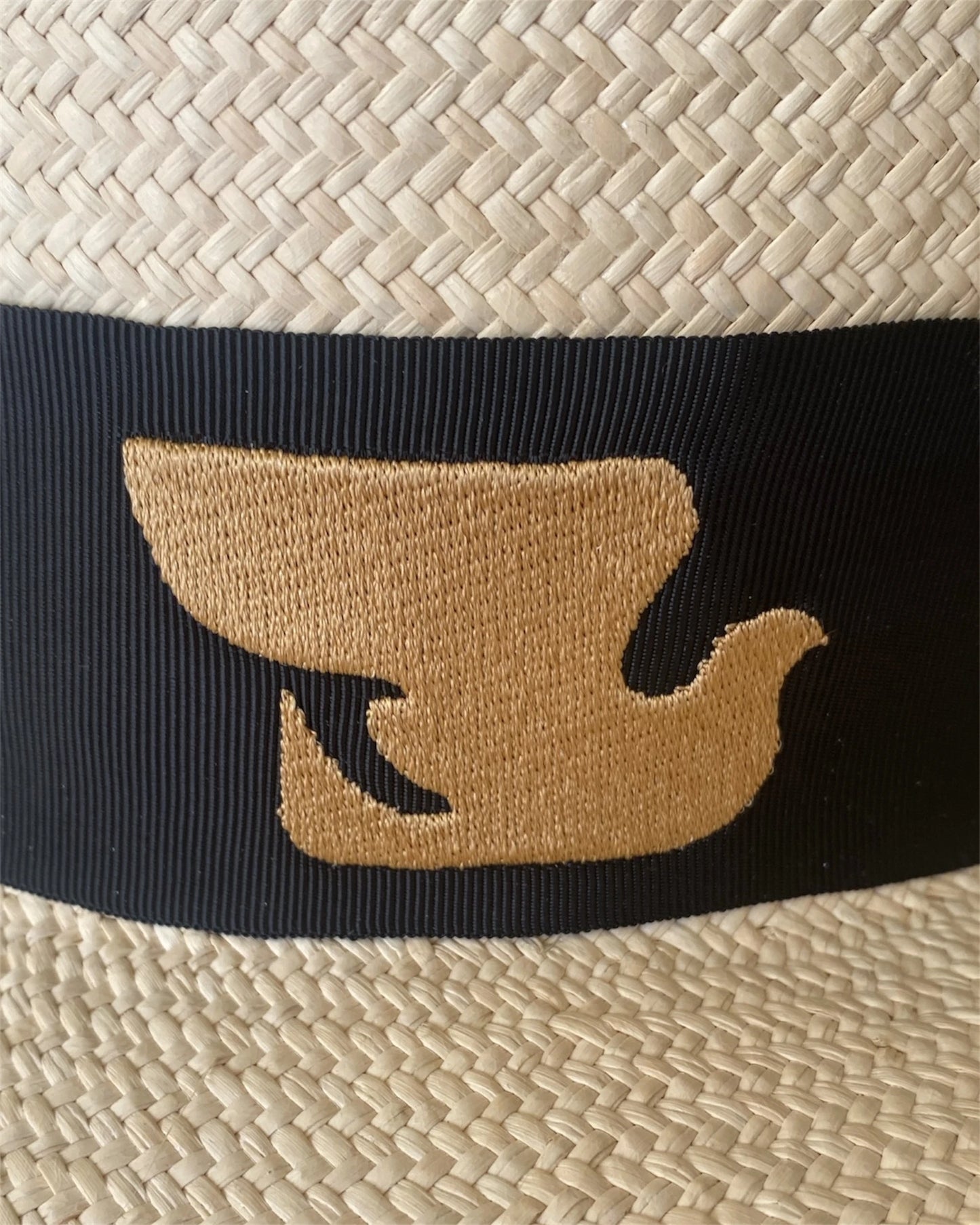 Detailed close-up of the Wayra Hat’s signature gold dove emblem embroidered onto a black grosgrain ribbon, set against finely handwoven natural straw for a timeless contrast.