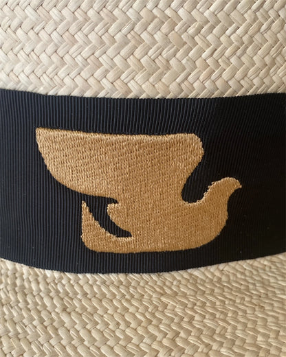 Detailed close-up of the Wayra Hat’s signature gold dove emblem embroidered onto a black grosgrain ribbon, set against finely handwoven natural straw for a timeless contrast.