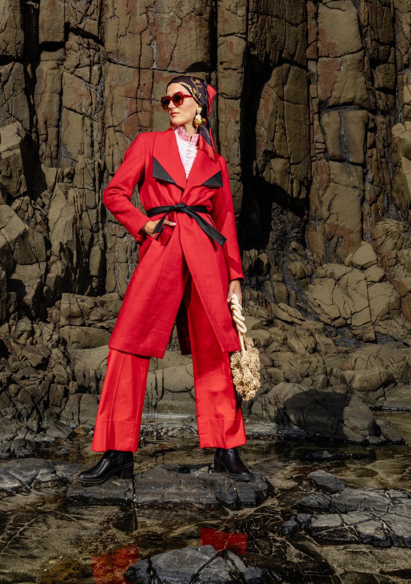 Front of Mia Belted Red Coat with black belt and sharp lapels, photographed in New York City – slow fashion elevated by My Paloma. Girl wearing the Mia Belted Taupe Coat with wide lapels and black accents, captured in Madrid, Spain – timeless tailoring by My Paloma.  Paired with a straw bag and a silk head scarf on a rocky shore