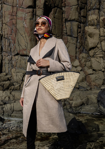 Girl wearing the Mia Belted Taupe Coat with wide lapels and black accents, captured in Madrid, Spain – timeless tailoring by My Paloma.  Paired with a straw bag and a silk head scarf on a rock shore