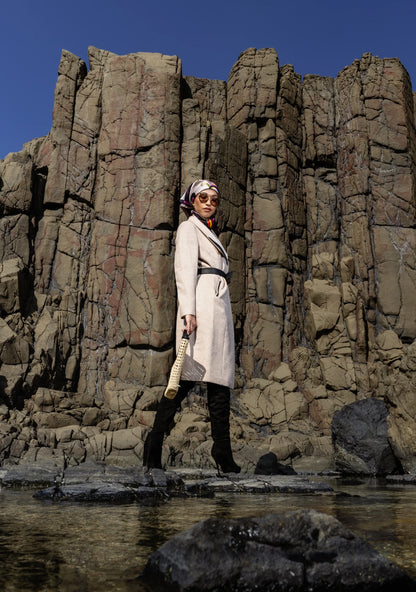 
Girl wearing the Mia Belted Taupe Coat with wide lapels and black accents, captured in Madrid, Spain – timeless tailoring by My Paloma.  Paired with a straw bag and a silk head scarf on a rock shore