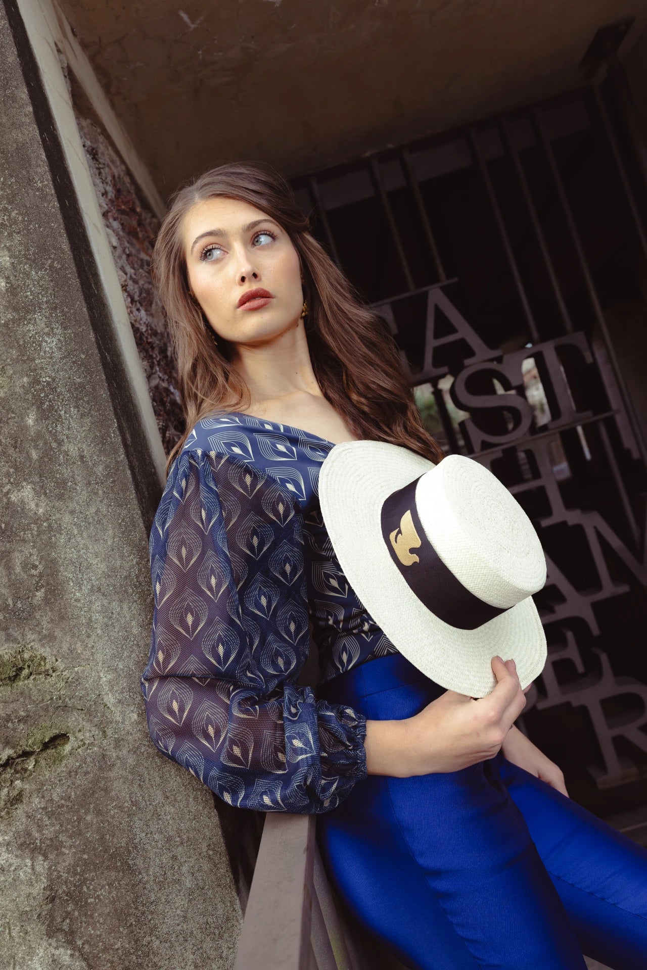 Model dressed in a navy patterned blouse and cobalt trousers, holding the Wayra Hat by her side—highlighting the hat’s structured crown, wide brim, and iconic dove ribbon band.