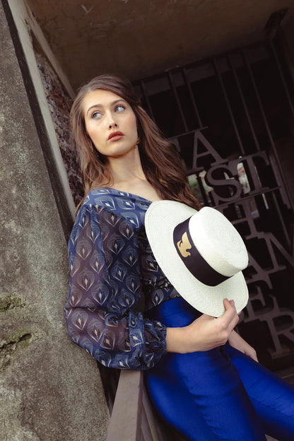 Model dressed in a navy patterned blouse and cobalt trousers, holding the Wayra Hat by her side—highlighting the hat’s structured crown, wide brim, and iconic dove ribbon band.