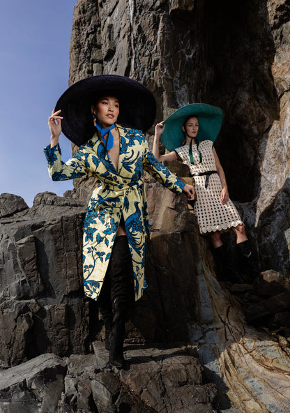 Model sposing in the Lady Ibiza Hat in navy blue and green, perched on rugged coastal rocks in an ocean-toned outfit—fusing sculptural millinery with windswept sophistication.