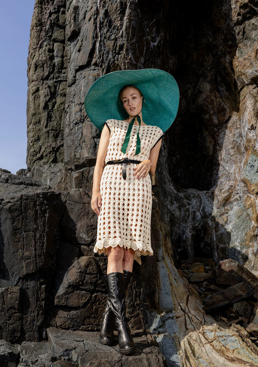 Model wearing the Lady Ibiza Hat in green, styled with a floral coat and black boots while standing against dramatic volcanic cliffs—blending boho elegance with bold natural backdrops.