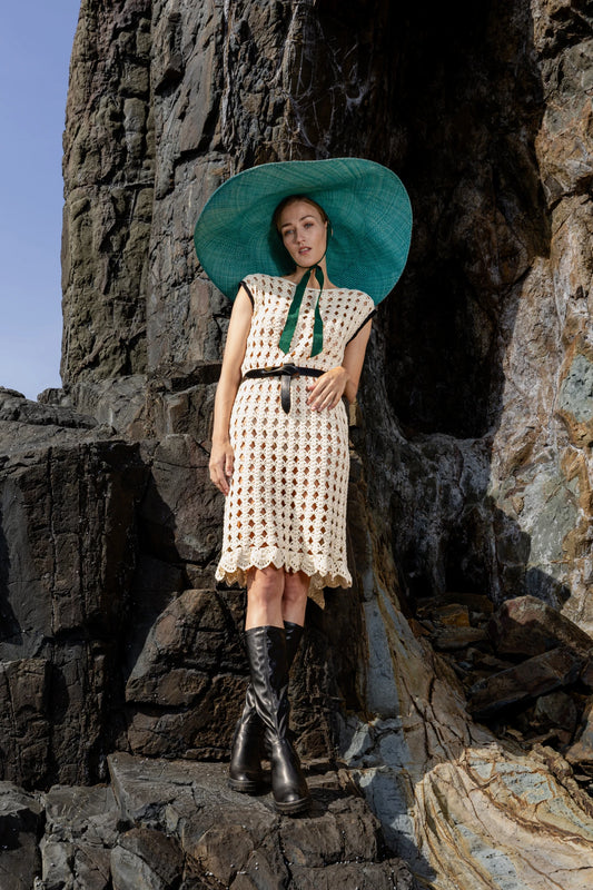Model seated on rocky coastline wearing the Mira hand-crochet dress paired with tall boots and a green wide-brimmed hat, exuding a bohemian coastal look.

