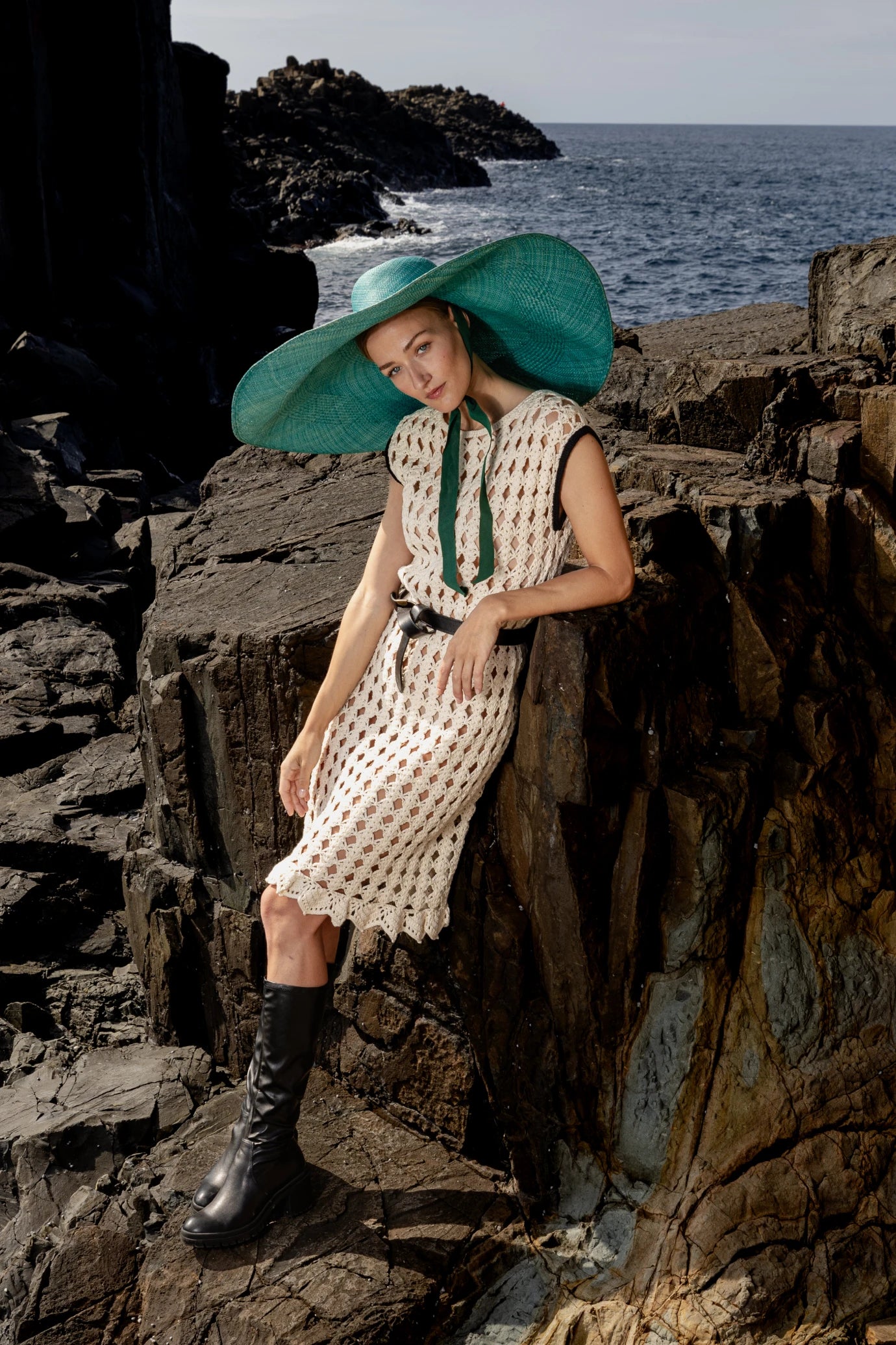 Model standing against natural rock formations in the Mira dress, styled with statement accessories and the signature wide-brimmed hat in green.