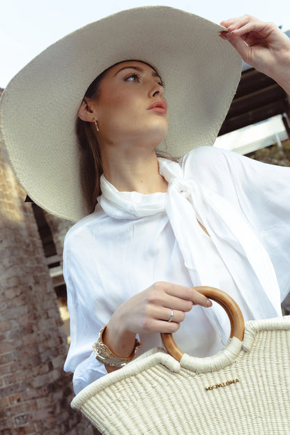 Close-up lifestyle shot of a model in a wide-brim hat holding the Verano Tote, capturing the tote’s rustic texture and polished circular handle in a relaxed, elegant mood.