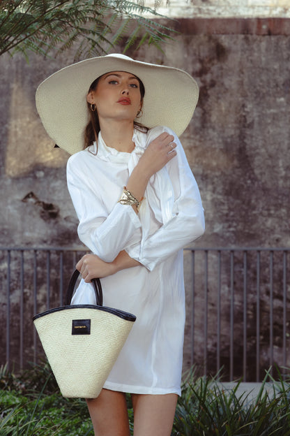 Model styling the Luna Hat with a white scarf-neck mini dress and bold gold cuff, standing in soft light under palm fronds—blending effortless glamour with Mediterranean inspiration.