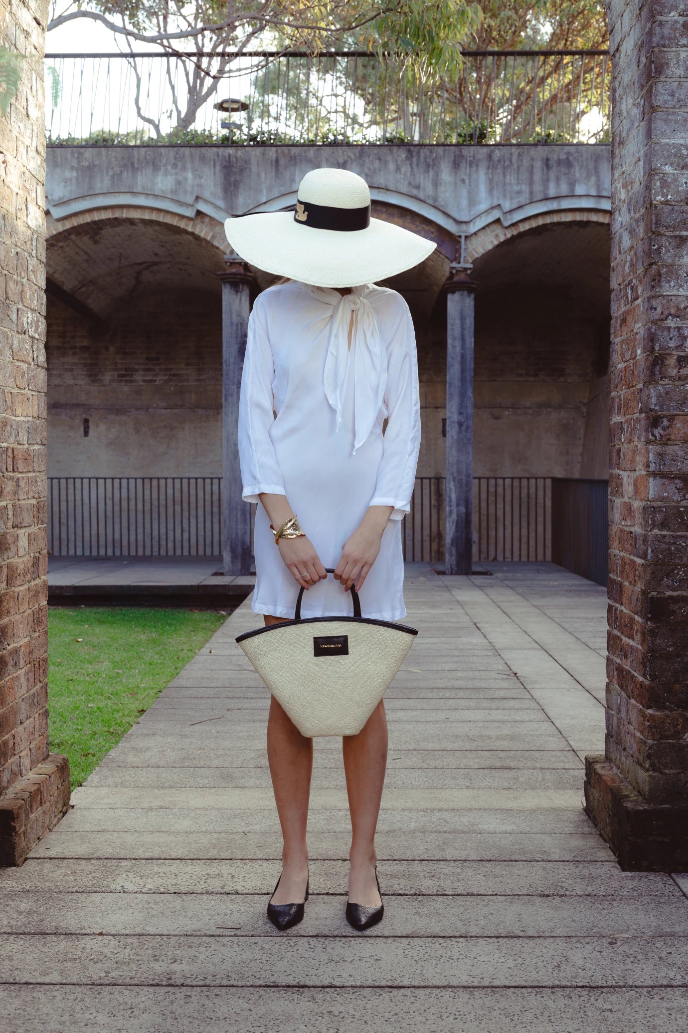 Full-body shot of model standing in an architectural archway with her head bowed, drawing full focus to the oversized Luna Hat and its dramatic silhouette—styled with monochromatic accessories for a Paris-meets-Riviera look.