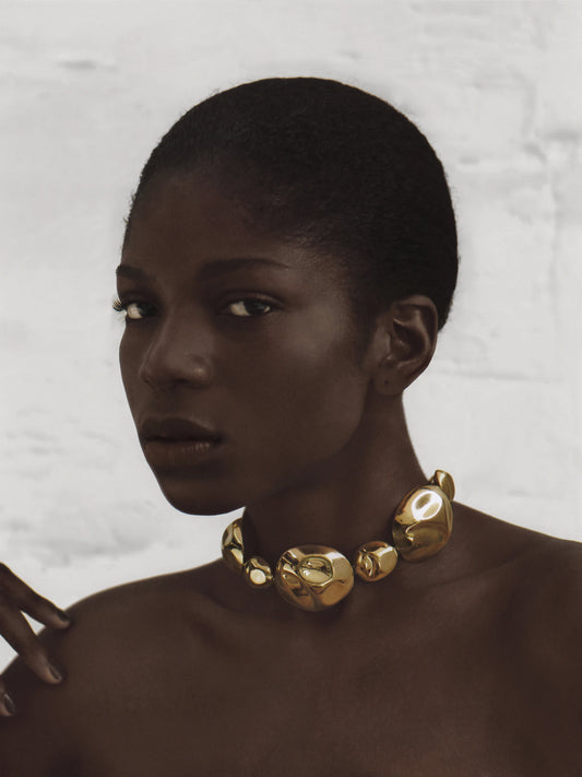 Dark-skinned model wearing the sculptural gold necklace with matching earrings, styled against a minimalist light backdrop.