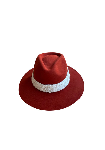 Flat lay of a brick red wool-felt fedora with a thick white embroidered band, highlighting the detailed crown and brim