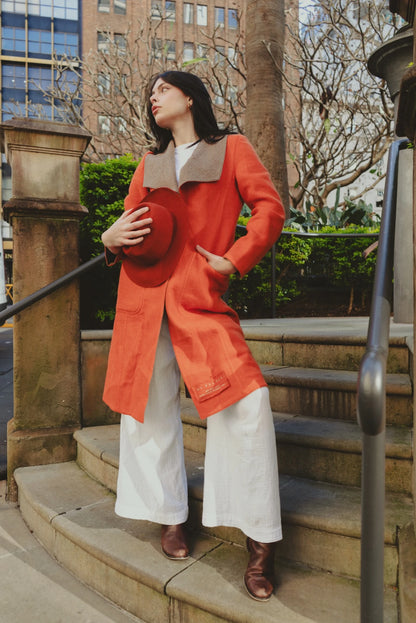 Stylish woman wearing a brick red wool-felt fedora and matching orange coat, standing near sandstone architecture in a city park.