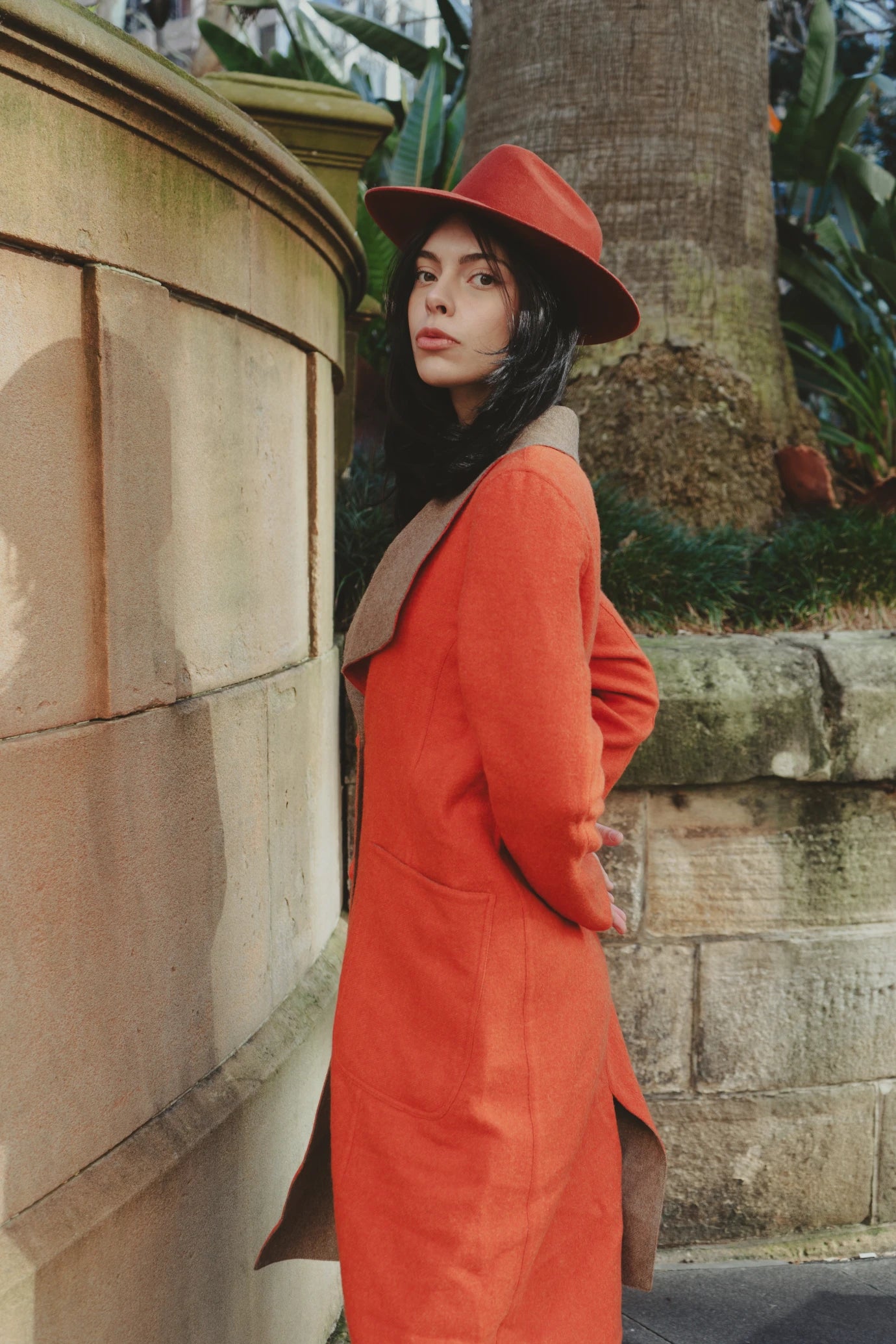 Woman in an orange overcoat and white trousers standing on stone steps, holding a brick red wool-felt fedora in an urban garden setting.