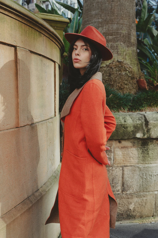 Woman in an orange overcoat and white trousers standing on stone steps, holding a brick red wool-felt fedora in an urban garden setting.