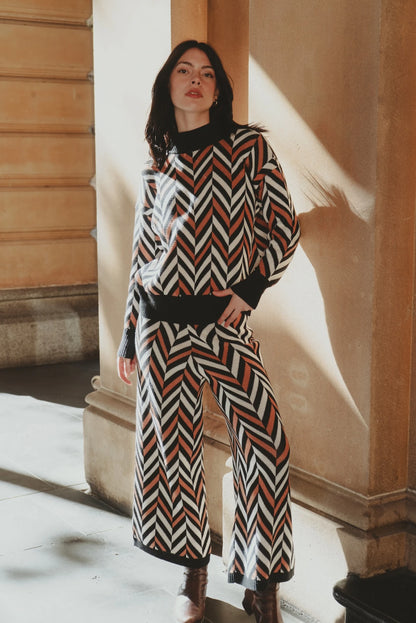 Model leaning against an arched stone pillar, wearing the My Paloma Zig Zag co-ord set with wide-leg pants and chevron print, styled with ankle boots for an elevated streetwear aesthetic in a luxury urban setting.