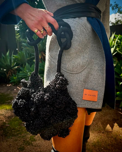 Model in blue printed dress holding the black Ameba Mesh Bag by the rocky cliffs of Lima’s coastline.